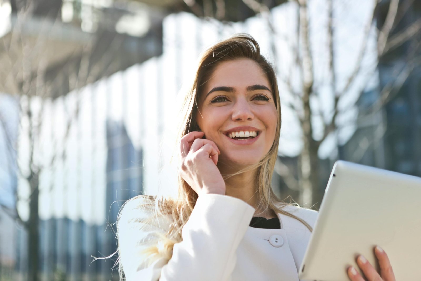 A lady talking on phone happily while holding a tablet outside